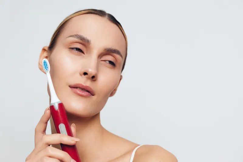 Young woman holding a red electric toothbrush near her face with closed eyes on a light background. Concept of oral hygiene, dental care, and personal grooming.
