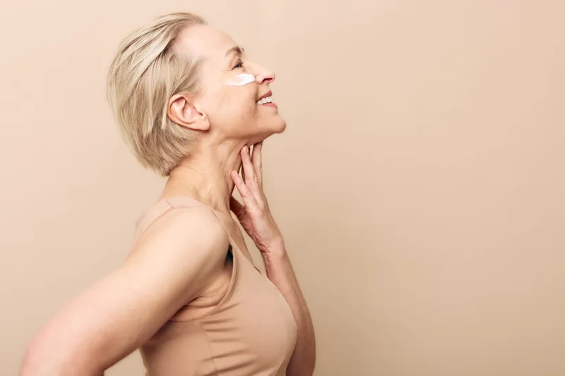 Smiling mature woman with short blonde hair applies white facial cream on cheeks, posing in beige top against soft beige background. Concept of skincare, self-care, wellness, and natural beauty.