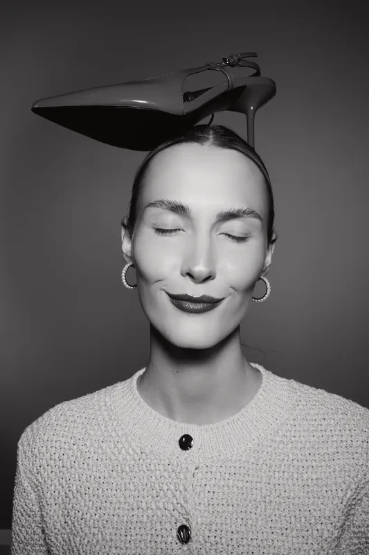 A smiling white woman with pale skin and closed eyes balances a high heel shoe on her head in a studio portrait concept of fashion and creativity.