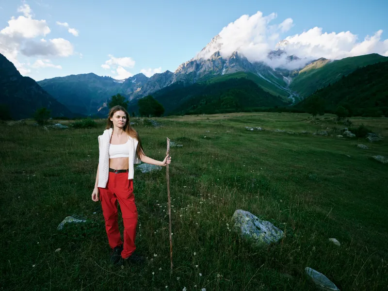 Woman in red pants holding a wooden staff stands in a lush meadow with distant mountains under a bright sky, capturing an expansive outdoor landscape and a sense of adventure