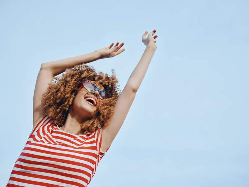 Joyful woman with curly hair wearing a red and white striped top raises arms under a bright blue sky, embodying carefree summer energy and confident, radiant mood.
