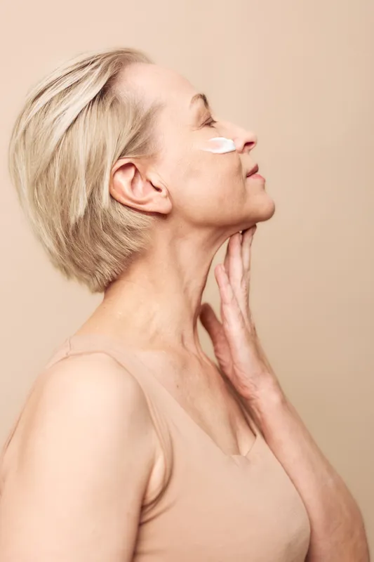 Profile of mature woman with short blonde hair applying cream on cheek, eyes closed, touching neck. Concept of skincare, aging skin care, beauty routine, self-care and wellness.