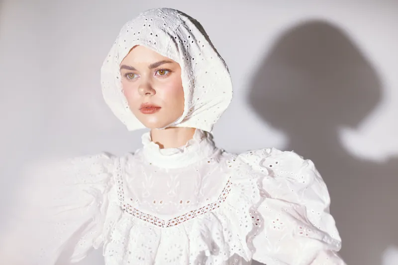 Ethereal female model in a white vintage dress with lace details and a matching headpiece, showcasing a serene expression against a soft, neutral background