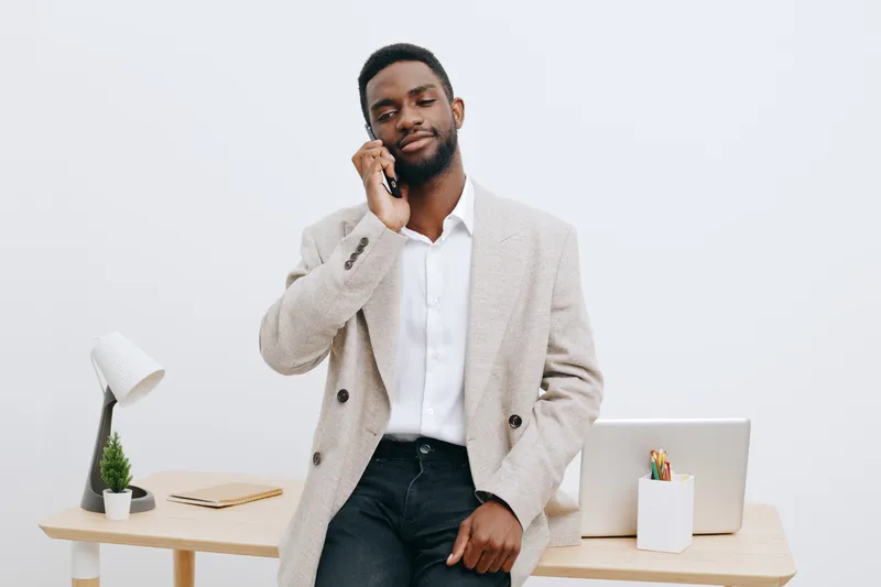 Confident young African American man in beige blazer and white shirt talking on mobile phone in modern office workspace with minimalistic design and wooden desk. People lifestyle concept