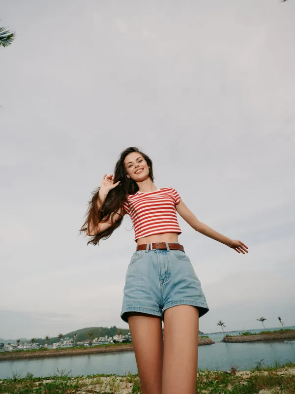 A young woman stands outdoors in casual summer attire, wearing a red and white striped crop top and denim shorts, smiling as wind lifts her hair.