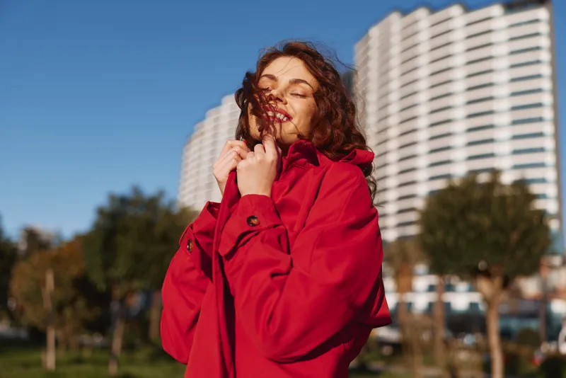 Smiling woman in a red coat stands outdoors with a cityscape backdrop, wind in her hair, sunny day, casual fashion, candid lifestyle portrait. Warm light enhances mood.