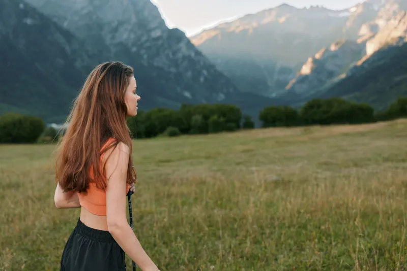 A lone woman strolls through a grassy meadow with distant mountains and a clear sky, creating a serene outdoor scene filled with quiet beauty and natural light.