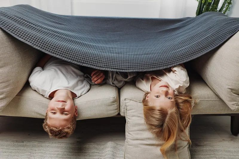Two children lie upside down on a sofa covered by a gray blanket. They look playful and cozy in a warm, light living room setting. Concept of family fun and childhood joy.