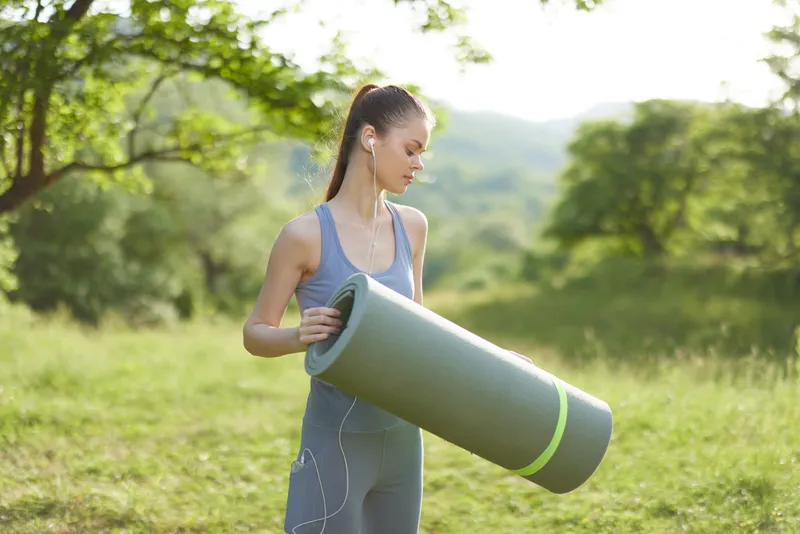 Young fitness female model in activewear holding yoga mat, enjoying outdoor exercise in nature during a sunny day