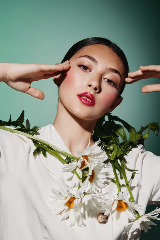 Elegant young woman with bright glossy lipstick and graphic eye makeup poses with daisies on white blouse against muted green background. Creative beauty and fashion concept.