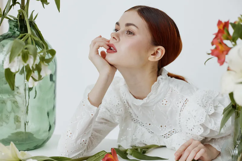 Thoughtful woman in a white vintage dress with red lipstick, surrounded by flowers, reflecting a serene and elegant mood