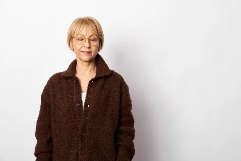 Senior woman with glasses wearing a brown coat, studio portrait on white background, calm neutral expression, fashion and age concept, soft lighting, looking at camera