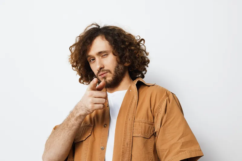 Confident young man with curly hair pointing at the camera, wearing a beige shirt, against a clean white background, showcasing a relaxed and stylish vibe