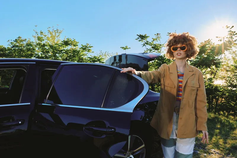 Curly haired woman in a beige jacket stands beside a dark car under bright sun, capturing a casual lifestyle with retro film color and warm, energetic mood