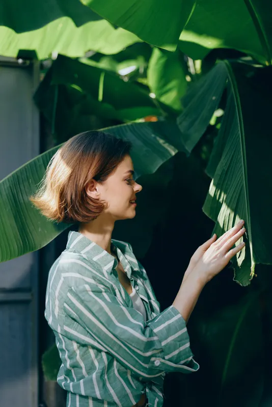 woman in striped shirt stands beside large green leaves, reaching to touch a tropical leaf, calm demeanor, soft light, peaceful moment in nature, a candid outdoor scene