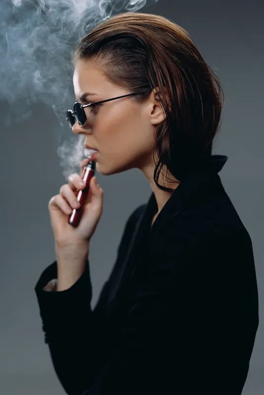 Stylish woman vaping with e-cigarette, dark background, focused expression, modern lifestyle concept.