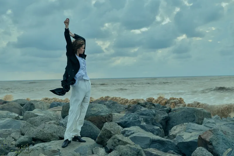 A woman stands on rugged rocks by the sea, arms raised in a victorious pose, facing a moody sky, conveying resilience, freedom and bold outdoor style, with wind tousling hair and distant surf.
