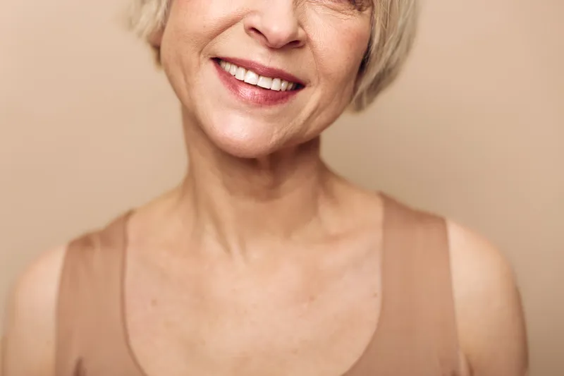 Close-up of smiling mature woman with short blonde hair and natural skin in beige tank top against soft beige background. Concept for happiness, aging, confidence, and natural beauty.