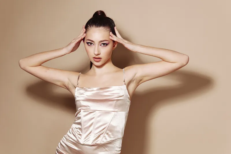 Elegant woman in a silky dress poses with hands on temples and intense gaze. Studio shot on a beige background. Creative beauty and fashion theme with striking makeup and style.