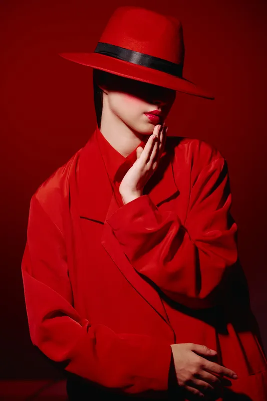 Stylish woman in red coat and wide hat covers eyes with shadow. Glossy red lips, soft hand pose. Studio shot with moody light for beauty, creative fashion concept.