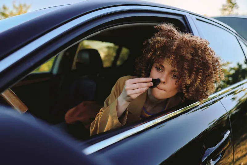 Young woman with curly hair sits in a car window, sniffing a small object, wearing a casual jacket, sunny day, candid suburban vibe.