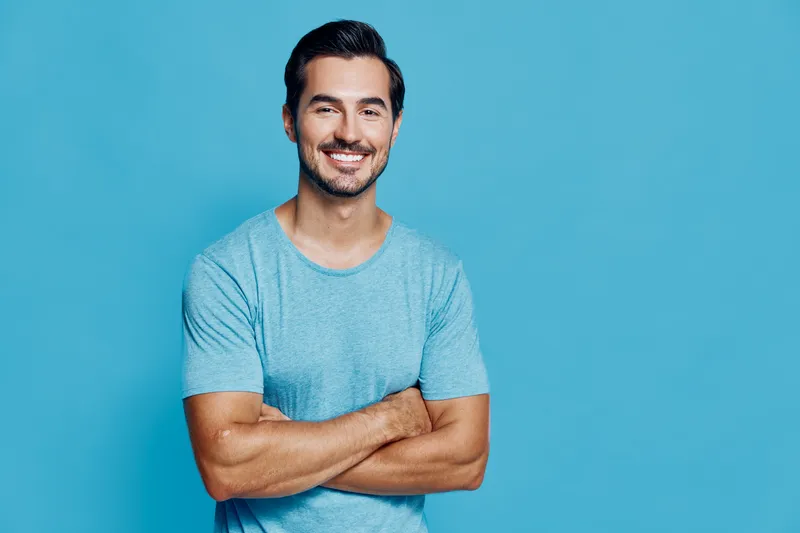 Smiling young man with crossed arms, wearing a light blue t-shirt, stands confidently in front of a solid blue background. Portrait captures joy and positivity.