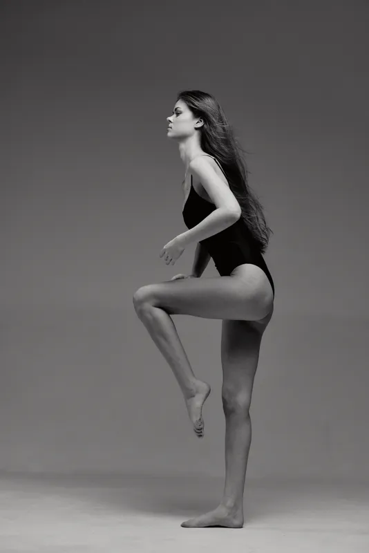 Black and white photo of a young woman in a leotard posing with raised leg, showing elegance, grace, and balance in studio lighting.