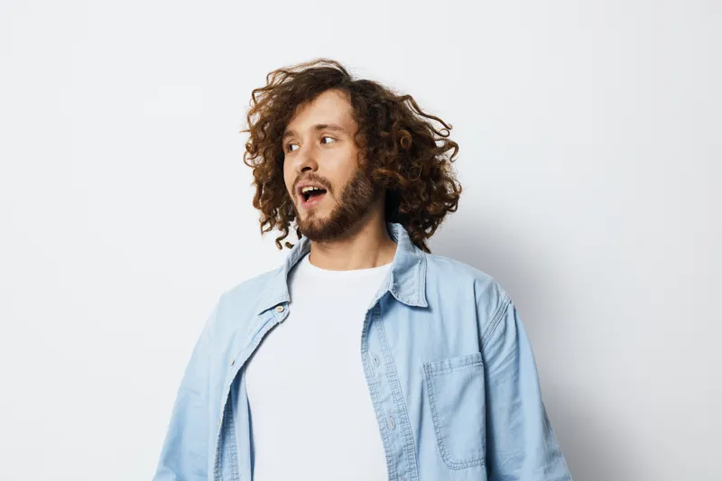 Cheerful young man with curly hair in casual attire, expressing joy against a minimalist light background, ideal for lifestyle and emotional wellness concepts