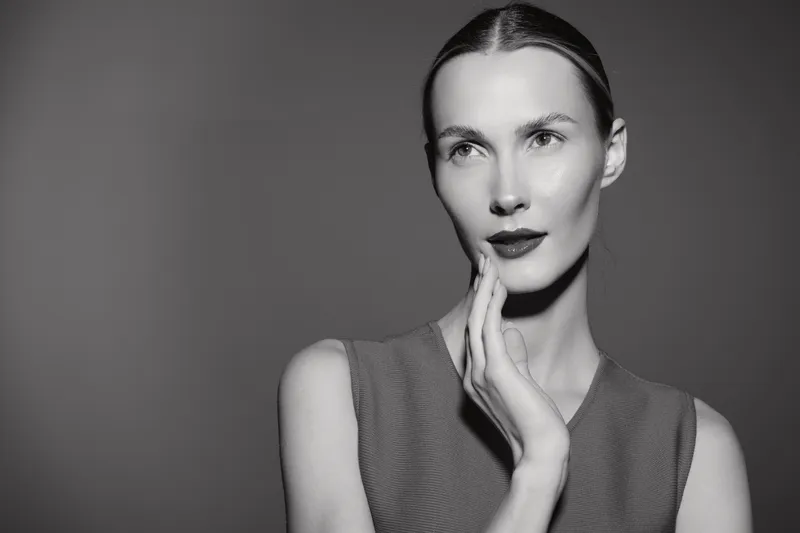 Portrait of a young Caucasian woman with fair skin and slicked back hair touching her face thoughtfully against a plain background. Concept of beauty, elegance, and contemplation.