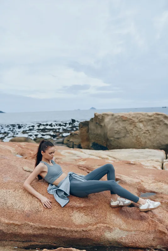 Fit young woman in athletic wear relaxing on rocky beach, showcasing active lifestyle and tranquility near the ocean in a calming color palette