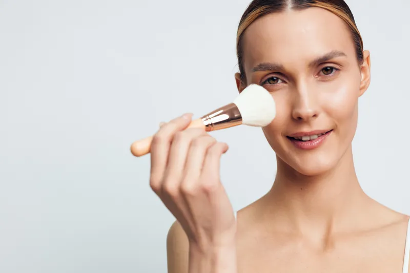 Young woman applying makeup with brush, smiling gently, clear skin, natural look, close-up portrait, light background, beauty routine, cosmetic brush in hand
