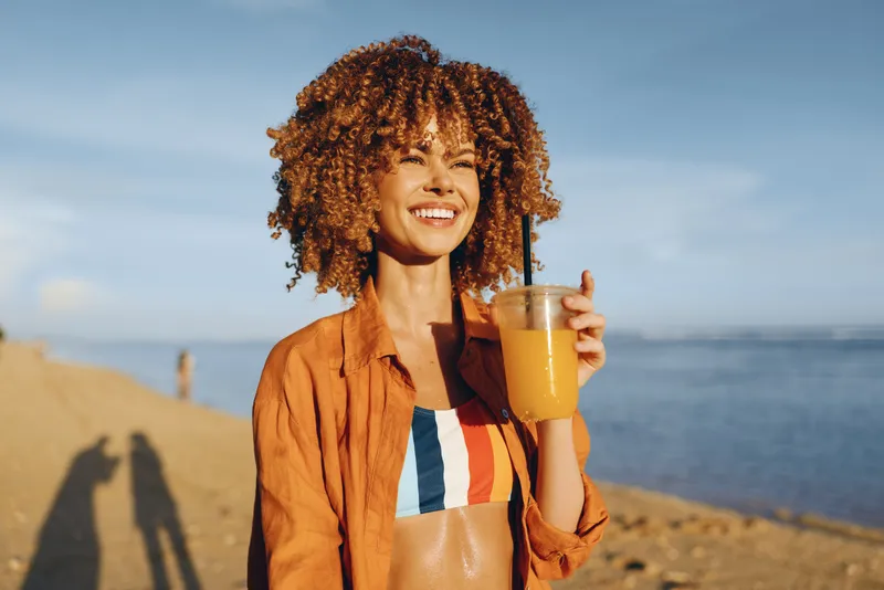 Woman curly hair sea drink sunshine summer smile outdoors portrait on the coast holding a glass of tea at golden hour warm light and calm breeze suggest relaxation and vacation travel
