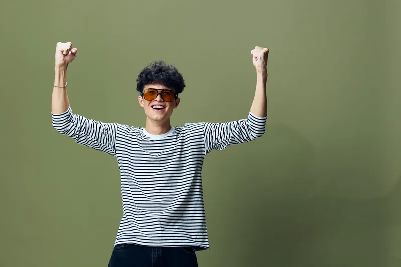 Young man celebrating success with raised fists, wearing striped shirt and sunglasses, joyful expression, isolated on green background, casual style