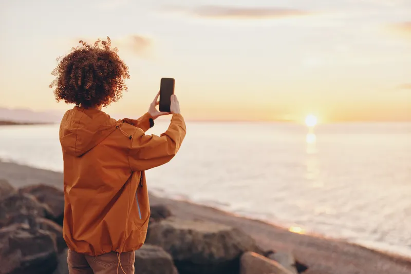 Woman lifestyle moment outdoors at sunset by the sea, curly hair in warm back view, holding a smartphone, orange jacket, calm relaxed travel mood, golden sky
