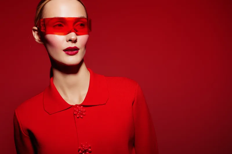 Fashion model in red outfit and red translucent glasses poses against red background. Concept of modern style, bold color, and high fashion photography with strong visual impact.