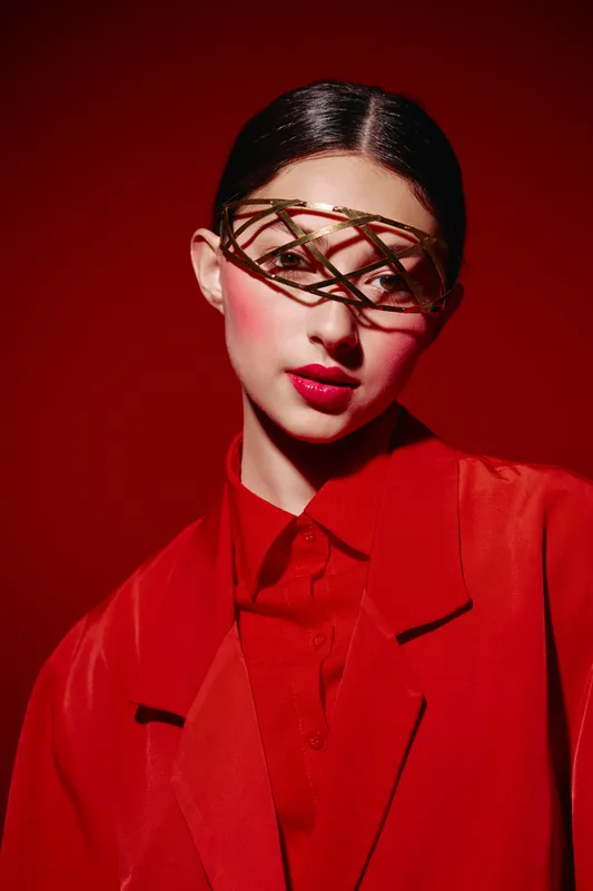 Young woman in avant-garde makeup and a golden unique glasses pose in a red oversized tuxedo against a deep red background. Beauty fashion creative studio portrait.