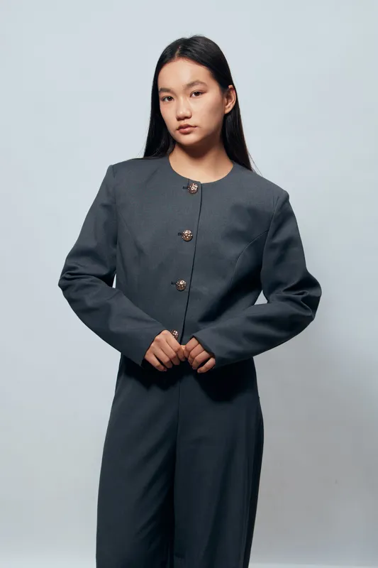Young woman standing with serious expression wearing modern dark gray suit jacket and pants against plain light background in studio for fashion and style portrait.