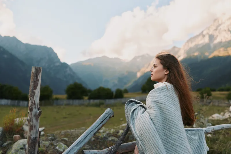 A woman wrapped in a gray knit blanket sits beside a weathered fence in a valley with distant mountains, conveying calm, solitude, and a connection with nature.