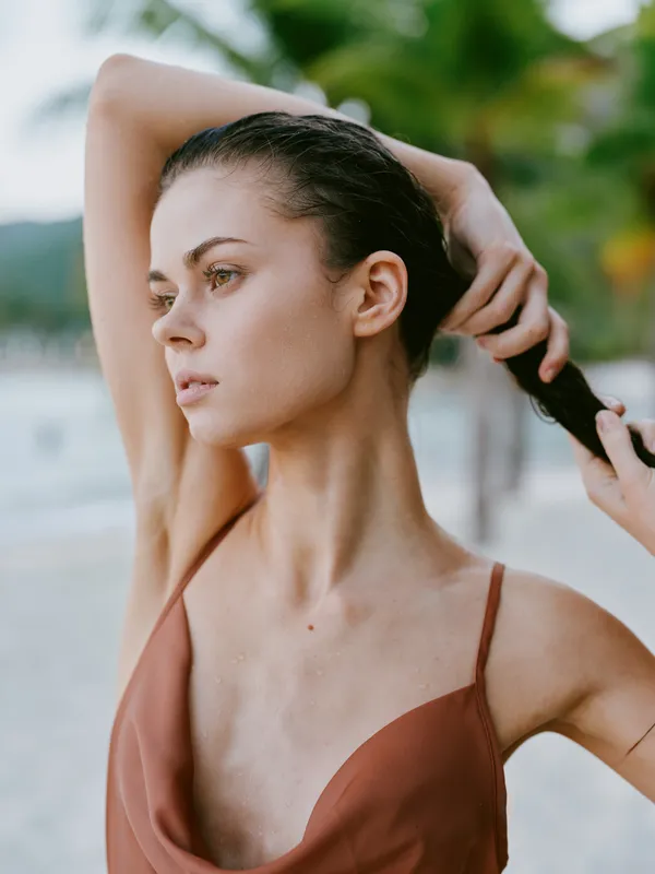 A portrait of a serene woman on a sunny beach, tying her wet hair back, wearing a rust brown swimsuit. Calm, focused expression under warm light.