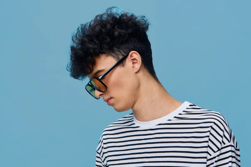 Young man with curly hair wearing black sunglasses and a striped shirt looking down against a pastel blue background.