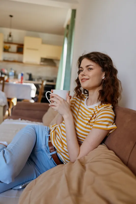 Casual woman sits on a couch at home, smiling while holding a mug in a sunlit living space, with a kitchen visible in the background. Cozy lifestyle moment, relaxed wellbeing vibe.