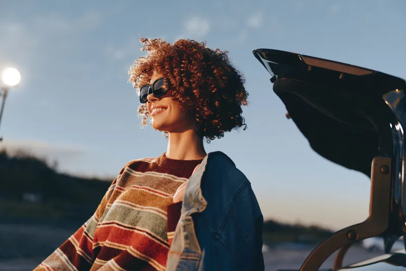 Woman lifestyle beside car at sunset, smiling with curly hair in denim jacket and striped sweater, outdoors on roadside, warm golden light, candid travel portrait.