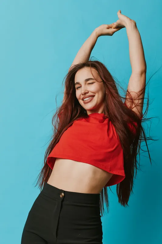 Young woman with long hair wearing red crop top and black pants stretching arms up with eyes closed and smiling against blue background for happiness and relaxation.