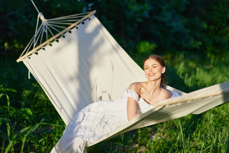 Young woman enjoying relaxation in a hammock on a sunny summer day, dressed in a light dress, smiling contently amidst a lush green background