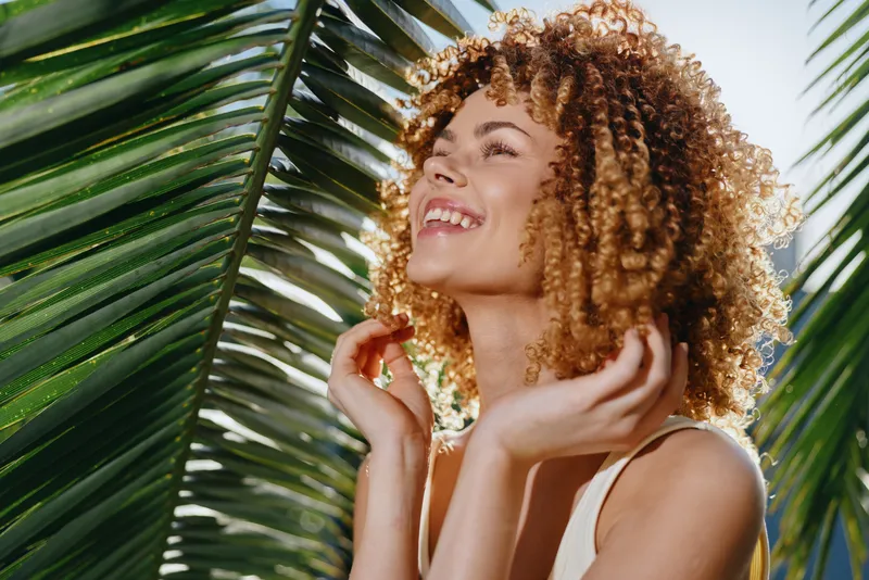 Smiling woman with curly hair enjoying sunlight outdoors, surrounded by lush green palm leaves, radiating happiness and natural beauty in warm daylight.