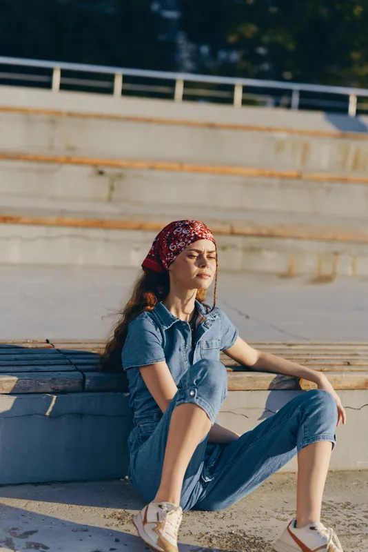 Modern boho-western denim street style woman sitting outdoors in film color, wearing a red headscarf and casual sneakers, enjoying natural sunlight and urban lifestyle.