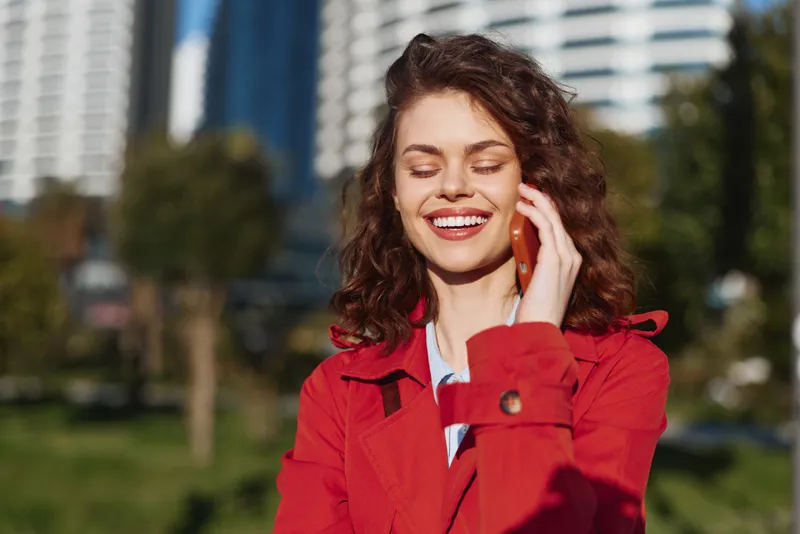 Smiling woman with curly hair wearing a red jacket chats on a smartphone in a sunny urban park with modern buildings and green grass nearby