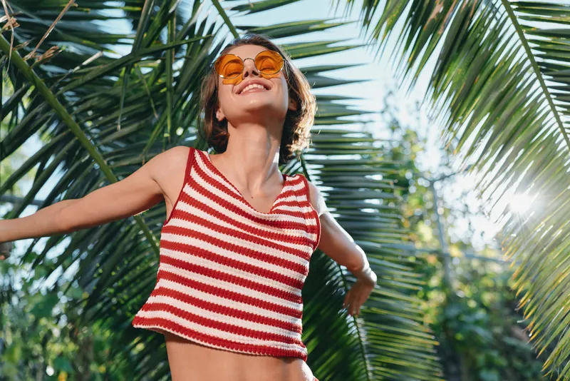 A cheerful woman stands outdoors in a sunny tropical setting, wearing a striped red and white top. She smiles with a carefree, vibrant mood, arms open, embracing warm breeze.