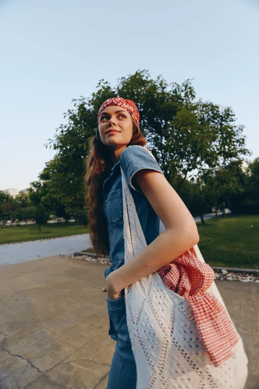 Lifestyle woman in modern boho-western denim street style standing outdoors with film color tones, red bandana, and casual tote bag looking confident and relaxed.