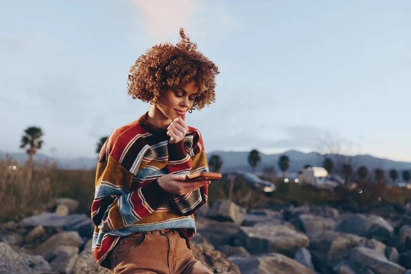 Curly hair woman in colorful sweater holds smartphone outdoor by rocky beach lifestyle scene thoughtful smile in natural dusk light everyday travel technology for calm communication